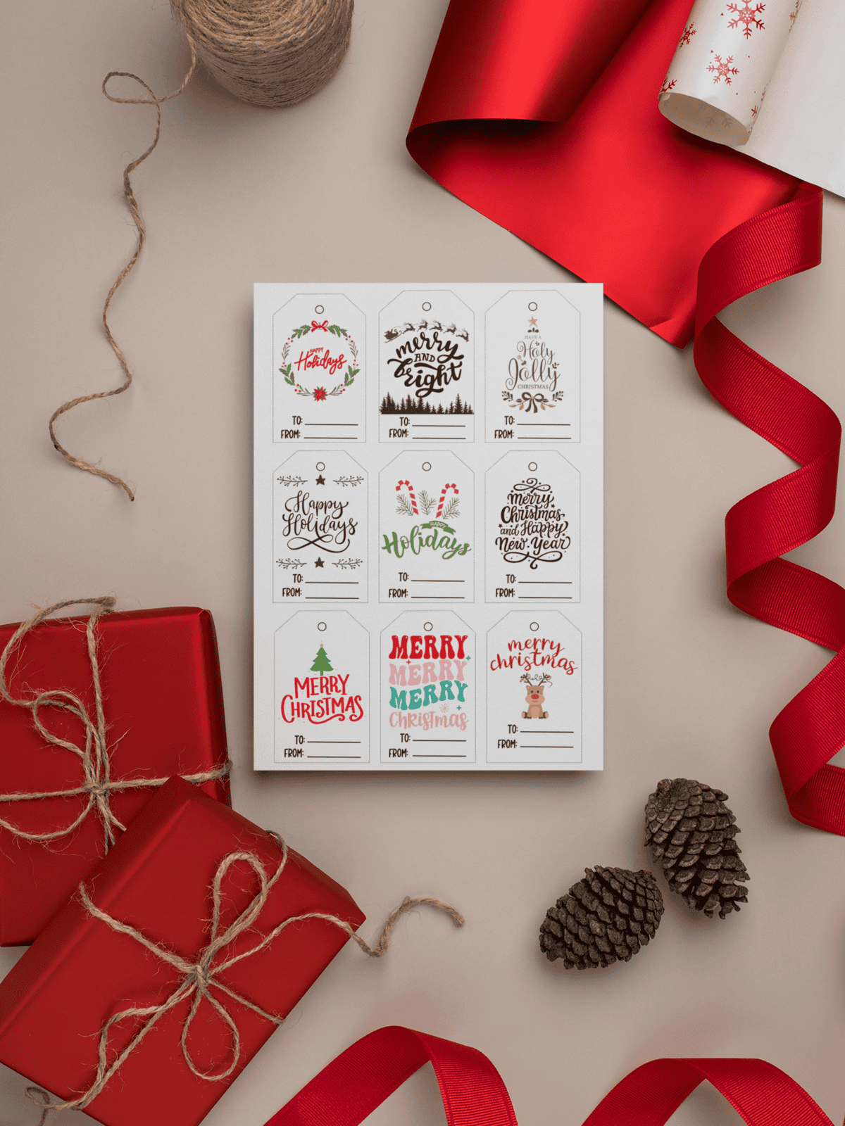 Set of printable Christmas gift tags featuring festive designs like wreaths, holiday greetings, and 'To/From' labels, surrounded by red wrapping paper, twine, and pinecones.